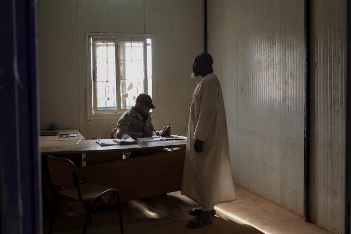 A border police officer sits at the desk and questions another man standing.