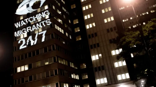 Office building at night with light projection showing watchful eye and reading "WATCHING MIGRANTS 24/7"