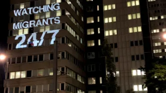 Light projection on UK Home Office building reading "WATCHING MIGRANTS 24/7"